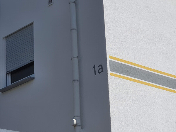 Das Bild zeigt einen vollendeten Fassadenanstrich an einem Wohnhaus. Zu sehen ist die sauber gearbeitete Hausnummer 1a, sowie ein farbliche Bordüre in gelb grau gelb.